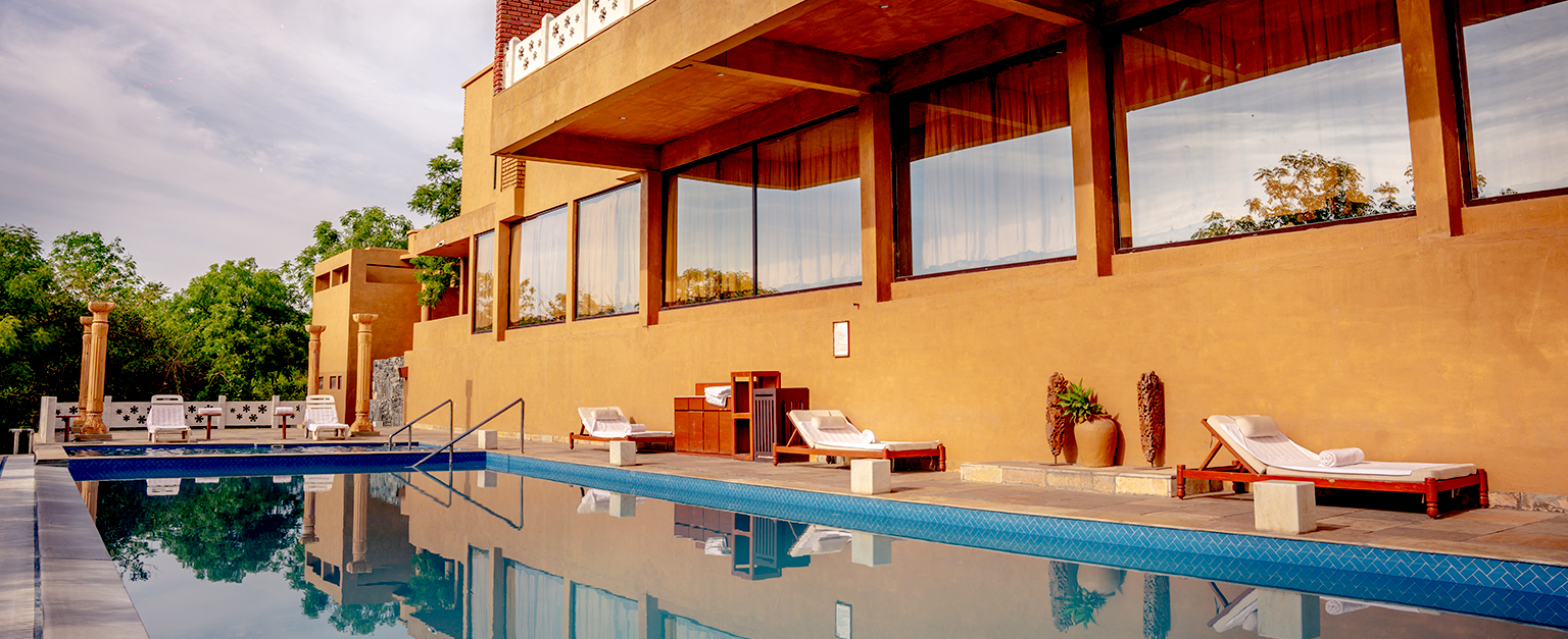 Outdoor pool area adjacent to a modern building with large windows, several white lounge chairs, and surrounding greenery.