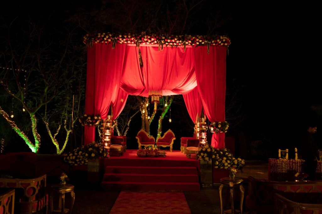 Experience the magic of a destination wedding with an outdoor stage at night adorned with red drapes, vibrant flower decorations, and enchanting illuminated surroundings.