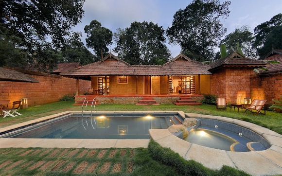 A quaint brick house with a tiled roof, surrounded by trees, features a lit swimming pool and adjacent hot tub in the foreground, reminiscent of the best destination wedding resorts in India, with deck chairs on a grassy area.