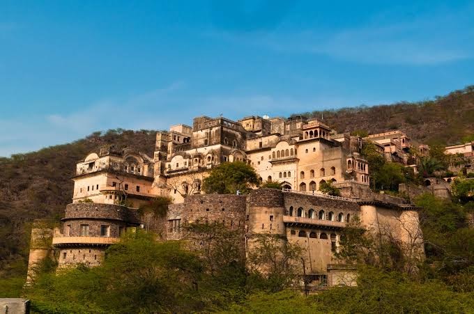 A large, historic fort complex with multiple structures and arches sits majestically on a hillside surrounded by trees under a clear blue sky, making it one of the best destination wedding resorts in India.