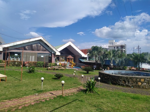 A beautifully landscaped garden with a stone fountain, bordered by neatly trimmed grass, sits in front of modern buildings under a partly cloudy sky—perfect for those seeking the best destination wedding resorts in India.