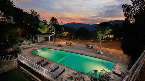 The outdoor swimming pool area at dusk, nestled among trees and hills, is reminiscent of the best destination wedding resorts in India. Streetlamps cast a warm glow over the lounge chairs as the sky bursts into a colorful sunset.