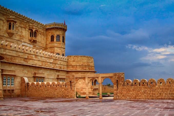 A historic sandstone fort with intricate architecture, featuring towers and arched windows, set against a cloudy sky, stands as one of the best destination wedding resorts in India.