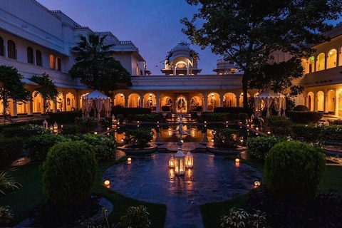 A dimly lit courtyard at dusk features a central path lined with lanterns, surrounded by lush greenery—a setting reminiscent of the best destination wedding resorts in India—with the building's illuminated arches in the background.