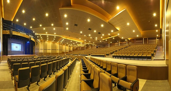 A large, empty auditorium reminiscent of the grand halls found in the best destination wedding resorts in India, featuring rows of brown seats under bright lights and a stage with a projector screen at the front.