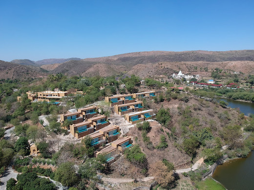 Aerial view of a hillside resort, one of the best destination wedding resorts in India, showcasing several buildings, each with a private pool. The surrounding landscape boasts green vegetation, a river, and distant hills under a clear blue sky.