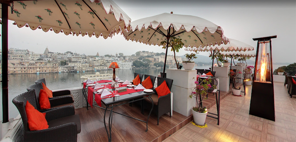 Discover one of the 17 best restaurants in Udaipur with a lake view. This rooftop gem features wicker chairs with red cushions, dining tables set for service, and offers breathtaking vistas of both the lake and cityscape, all under elegant white umbrellas with heaters nearby.